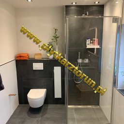 Paweł Kowal - Nowoczesna łazienka z ciemnymi płytkami, kabiną prysznicową ze szklanymi drzwiami, wiszącą toaletą i dekoracyjnymi detalami: bambus w wazonie, pomarańczowe ręczniki i drewniane akcenty.