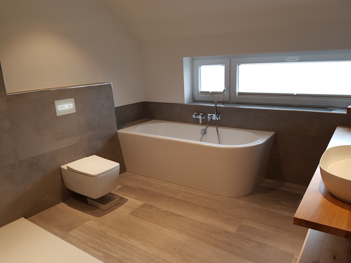Nowoczesna łazienka po remoncie: wolnostojąca wanna, podwieszana toaleta z ukrytym systemem spłukiwania, drewnopodobne płytki podłogowe, szare płytki ścienne i okno z roletą.