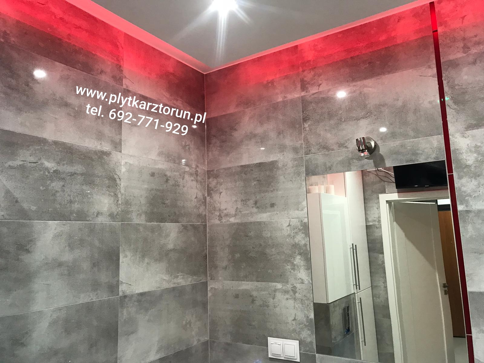 Wykończona łazienka z szarymi płytkami imitującymi beton, oświetleniem LED w kolorze czerwonym i lustrem z odbiciem szafki oraz fragmentu drzwi. Na płytkach widoczny adres strony internetowej...