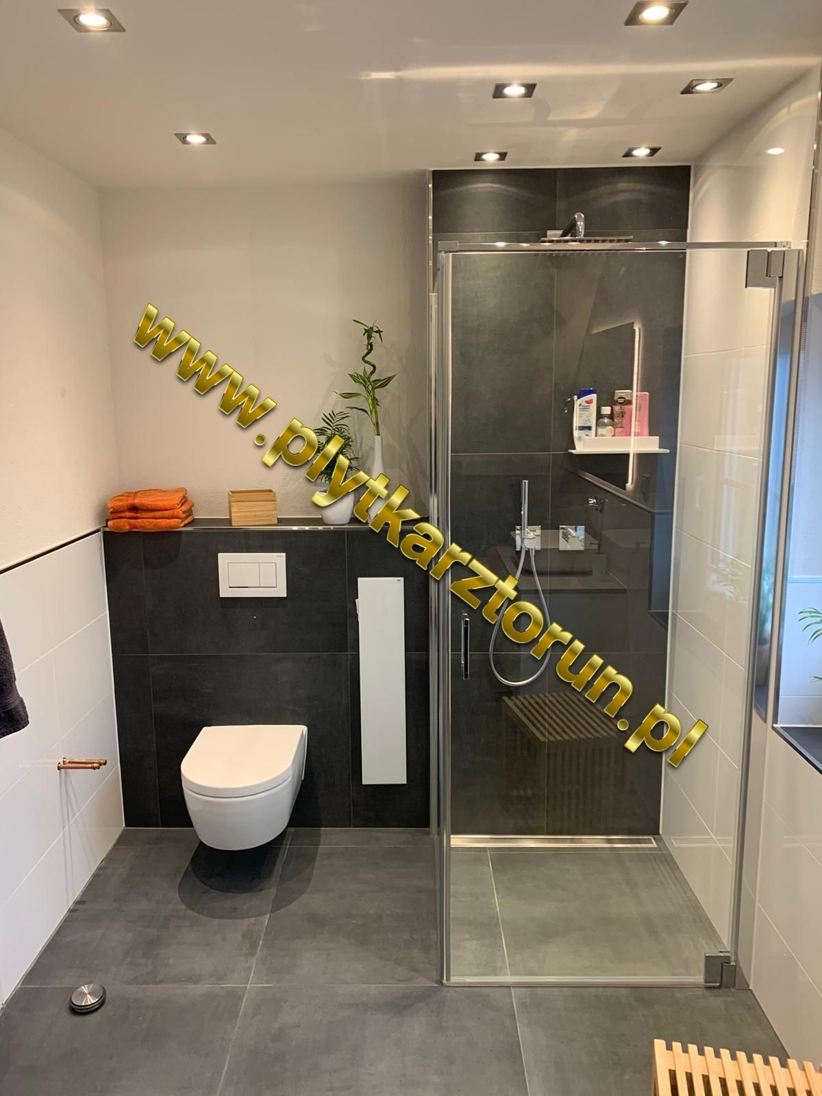 Nowoczesna łazienka z ciemnymi płytkami, kabiną prysznicową ze szklanymi drzwiami, wiszącą toaletą i dekoracyjnymi detalami: bambus w wazonie, pomarańczowe ręczniki i drewniane akcenty.