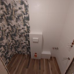 Kompleksowe Wykończenie Wnętrz Grzegorz Dewicki - Wnętrze łazienki w trakcie remontu: białe płytki na ścianach, jedna ściana z motywem liści, drewnopodobne panele na podłodze, podwieszana toaleta w zabudowie, wystające kable elektryczne i rury...