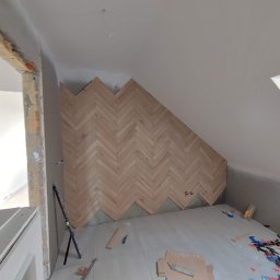 Kompleksowe Wykończenie Wnętrz Grzegorz Dewicki - Montaż drewnianej okładziny ściennej w jodełkę na poddaszu w Poznaniu, widoczne niedokończone fragmenty i narzędzia glazurnicze na podłodze.