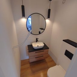 Kompleksowe Wykończenie Wnętrz Grzegorz Dewicki - Nowoczesna łazienka z drewnianą szafką pod umywalką, okrągłym lustrem z czarną ramą i minimalistycznymi lampami wiszącymi, białe ściany z delikatną teksturą i drewnopodobna podłoga, widoczne rury...