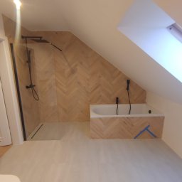 Kompleksowe Wykończenie Wnętrz Grzegorz Dewicki - Wykończona łazienka na poddaszu ze skośnym oknem, kabiną prysznicową typu walk-in z czarną armaturą i wanną obudowaną płytkami ułożonymi w jodełkę, zabezpieczona niebieską taśmą malarską.