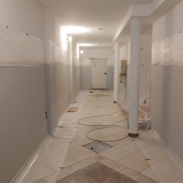 Kompleksowe Wykończenie Wnętrz Grzegorz Dewicki - Korytarz biurowy w trakcie przygotowań do malowania, ściany zabezpieczone folią malarską, widoczne kolumny i przewody na podłodze.