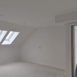 Kompleksowe Wykończenie Wnętrz Grzegorz Dewicki - Poddasze po szpachlowaniu i malowaniu na biało, z trzema oknami dachowymi i widocznym przewodem elektrycznym zwisającym z sufitu, przez otwarte drzwi balkonowe widać plac budowy.