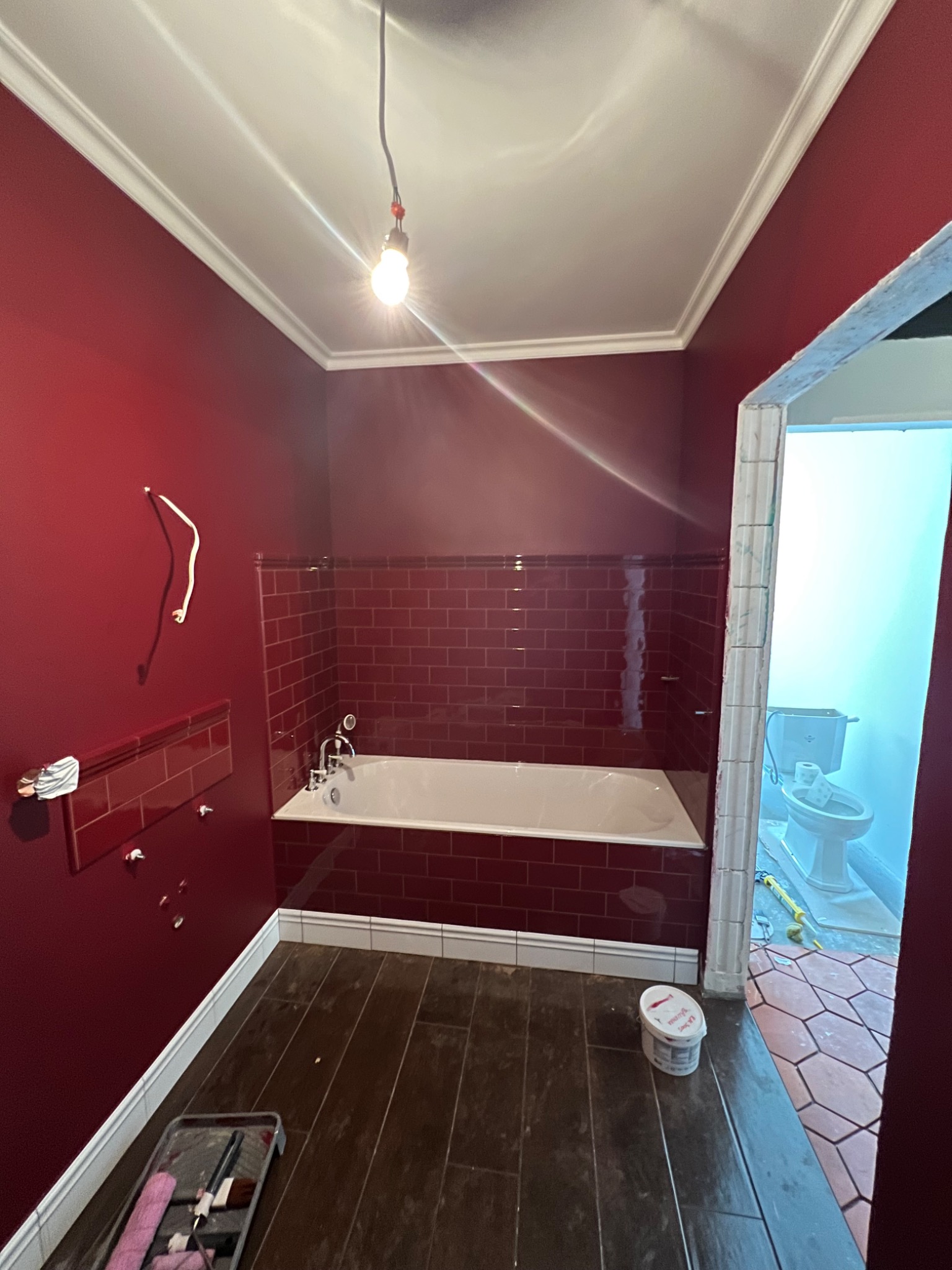 Remontowana łazienka z bordowymi ścianami i płytkami, widoczna wanna, ciemna podłoga, akcesoria malarskie i przejście do toalety z terakotą na podłodze.