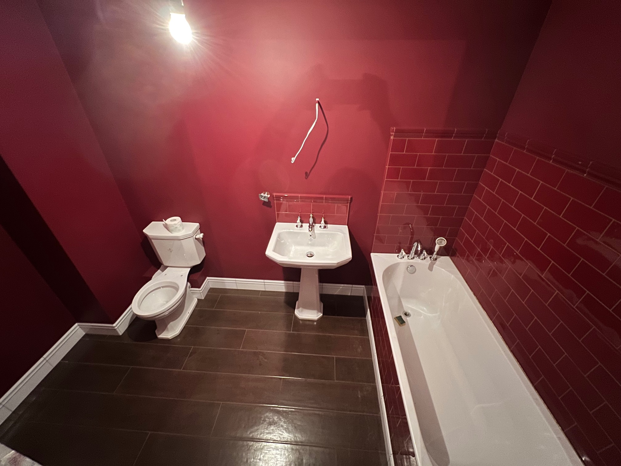 Czerwona łazienka z ciemnobrązową podłogą z płytek imitujących drewno, białą ceramiką sanitarną i częściowo wykończona czerwoną glazurą.