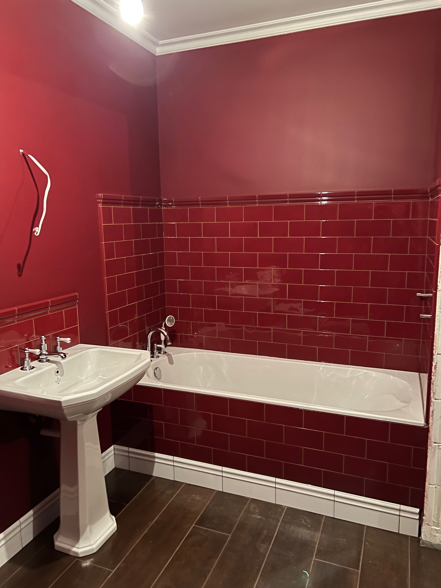 Czerwona łazienka z białą wanną i umywalką na postumencie, ściany wyłożone błyszczącymi, czerwonymi płytkami w stylu retro z jasną fugą, ciemna drewnopodobna podłoga, widoczne niedokończone prace...