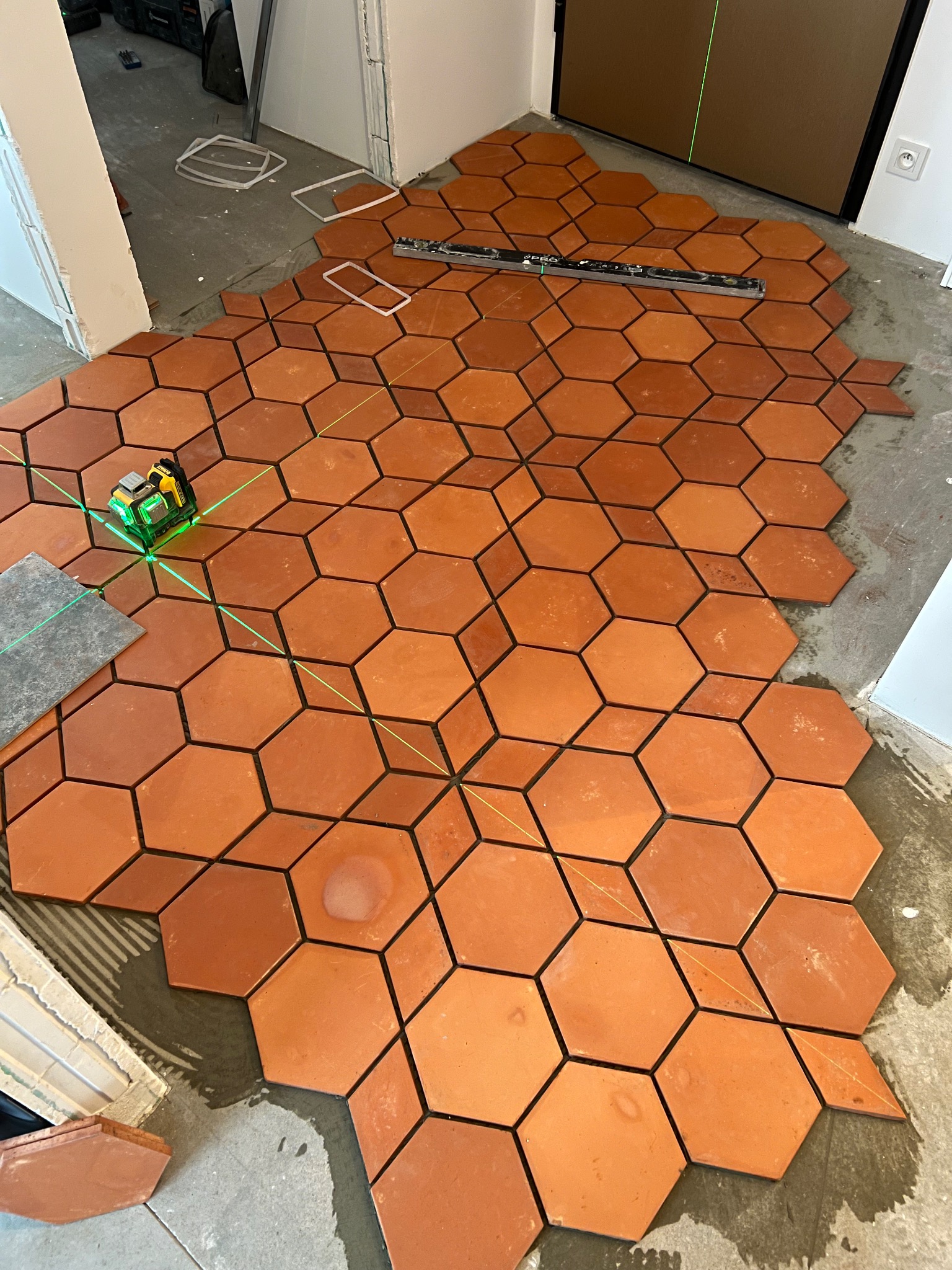 Ułożone w części pomarańczowe, heksagonalne płytki podłogowe z fugami w kształcie rombów, poziomica i laser krzyżowy wyznaczają linie proste, widoczny fragment podłoża i niezakończone krawędzie.