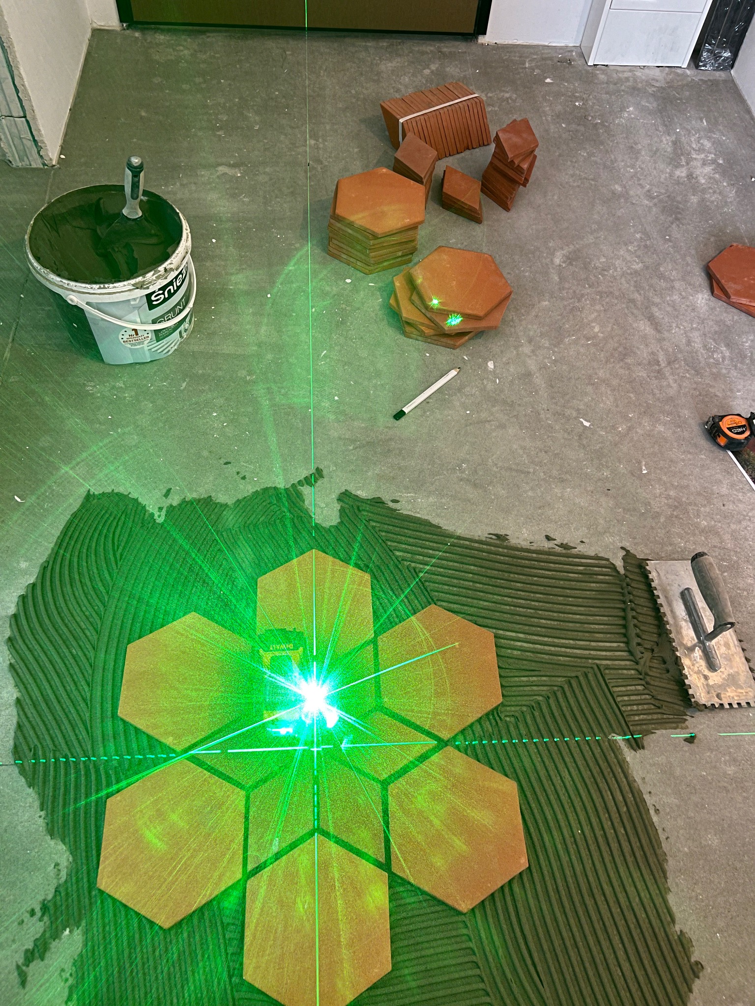 Układanie heksagonalnych płytek podłogowych z użyciem lasera krzyżowego, widoczny klej na podłodze, szpachelka z pacą, wiadro z gruntem Sniezka oraz ułożone płytki w tle.