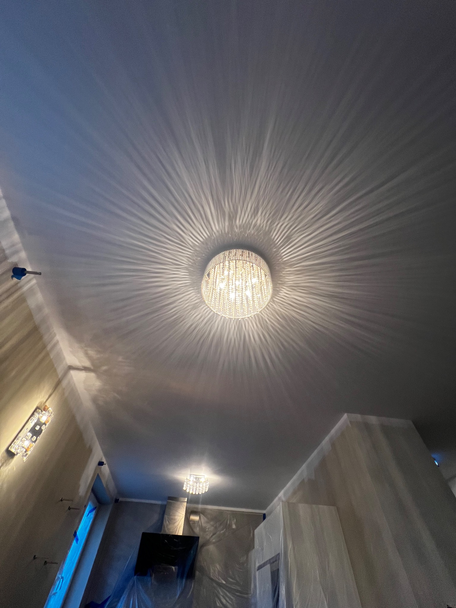 Kryształowy żyrandol na suficie w pokoju zabezpieczonym folią malarską, widoczne ślady po taśmie malarskiej na ścianach, przygotowanie do malowania.