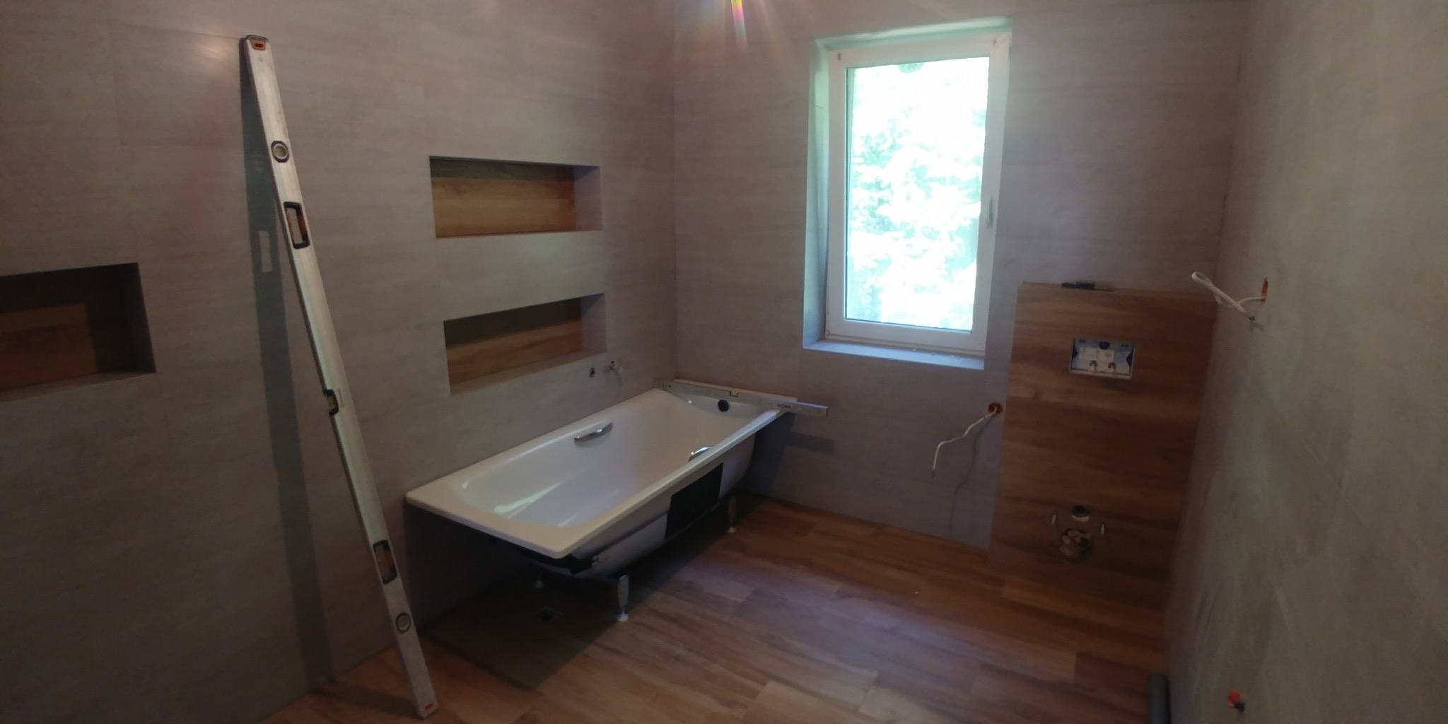 Łazienka w trakcie remontu z wanną na nóżkach, poziomica oparta o ścianę z wnękami, drewnopodobne płytki podłogowe i fragment ściany wyłożony drewnem.