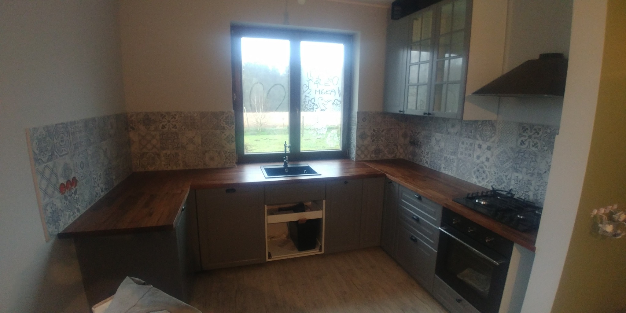 Wykończona kuchnia z szarymi szafkami, drewnianym blatem i dekoracyjnymi płytkami na ścianie, widok na zewnątrz przez okno z rysunkami.