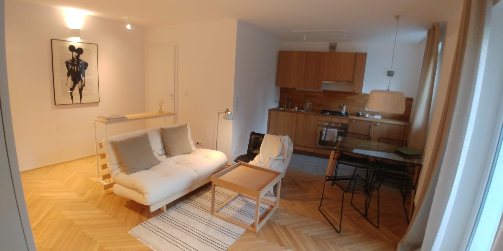 Jasne wnętrze mieszkania z parkietem w jodełkę, sofą z beżowymi poduszkami, drewnianym stolikiem kawowym i otwartą kuchnią z drewnianymi szafkami.