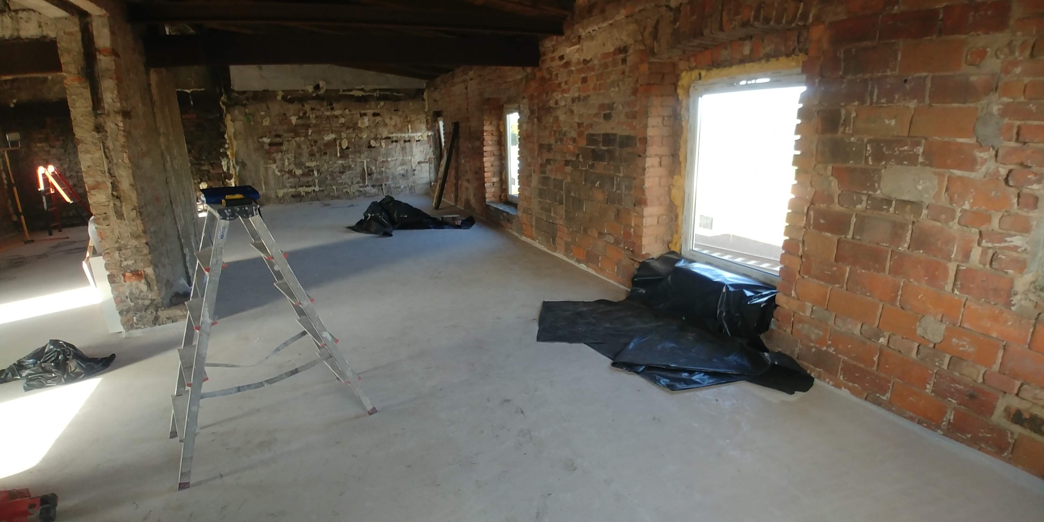 Wnętrze w trakcie remontu: surowe ceglane ściany, świeżo wylana podłoga, zabezpieczone okna folią, drabina aluminiowa i lampa budowlana w tle.