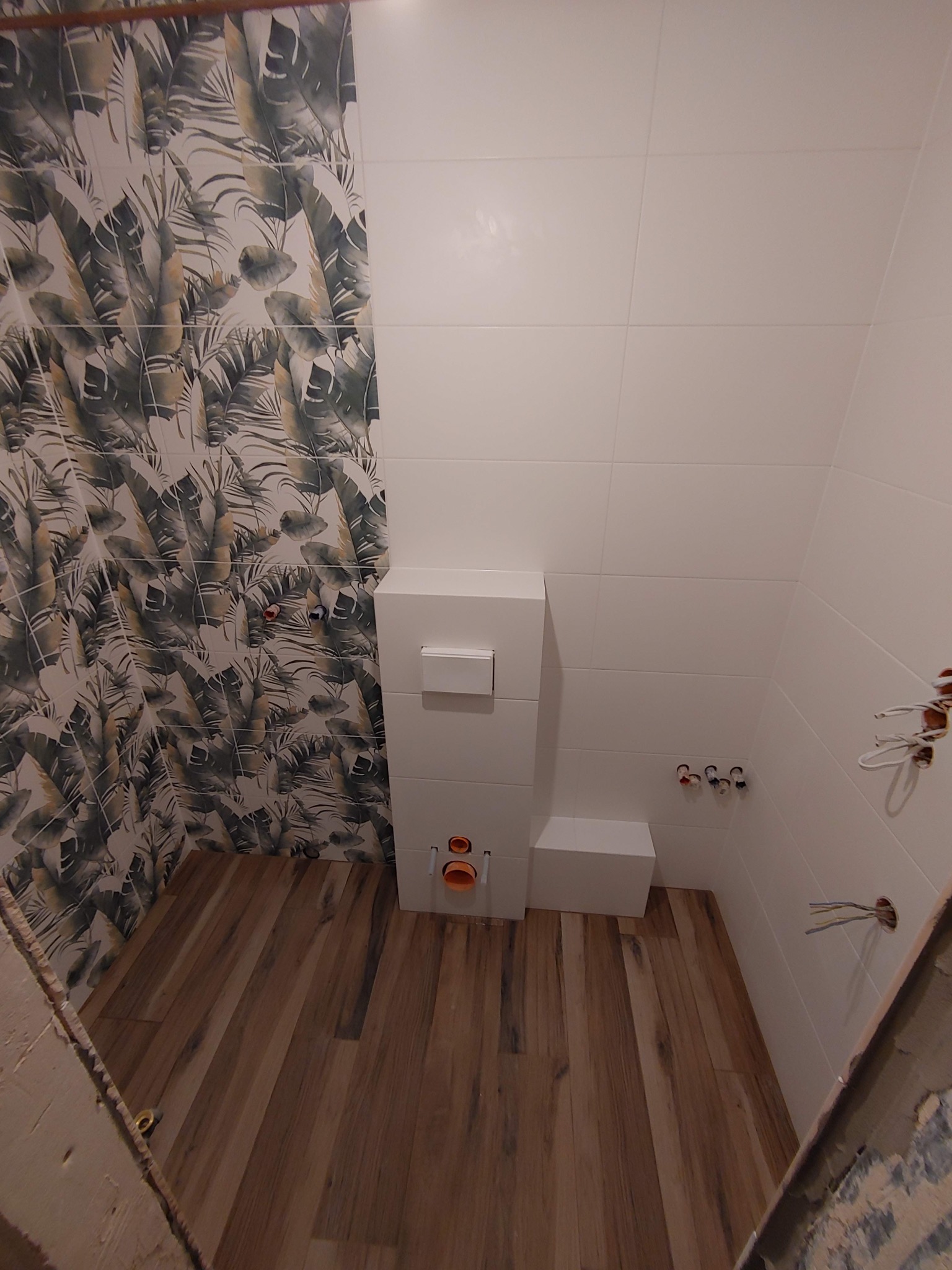 Wnętrze łazienki w trakcie remontu: białe płytki na ścianach, jedna ściana z motywem liści, drewnopodobne panele na podłodze, podwieszana toaleta w zabudowie, wystające kable elektryczne i rury...