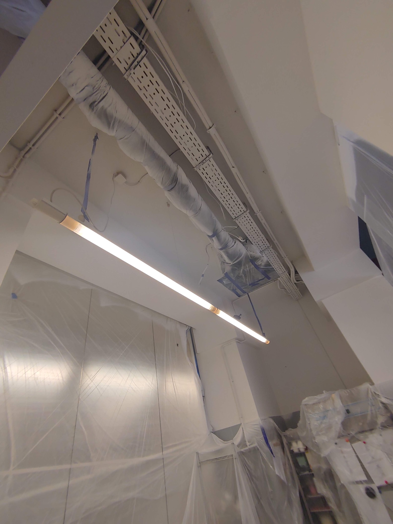 Przygotowania do prac malarskich w biurze: zabezpieczone folią ściany, sufit z instalacjami owiniętymi folią ochronną i oświetlenie liniowe.