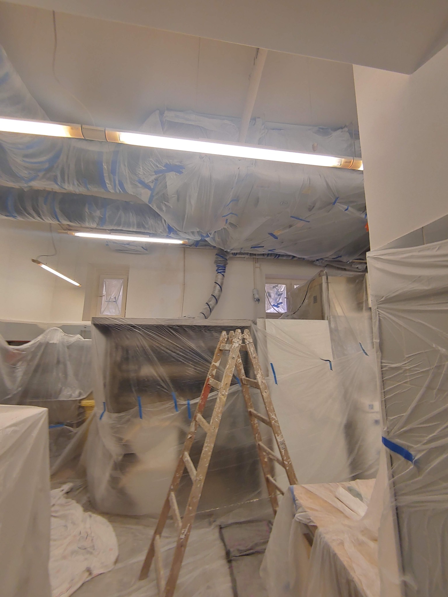 Przygotowania do prac wykończeniowych w pomieszczeniu biurowym, meble i elementy wyposażenia zabezpieczone folią malarską, widoczna drabina malarska.