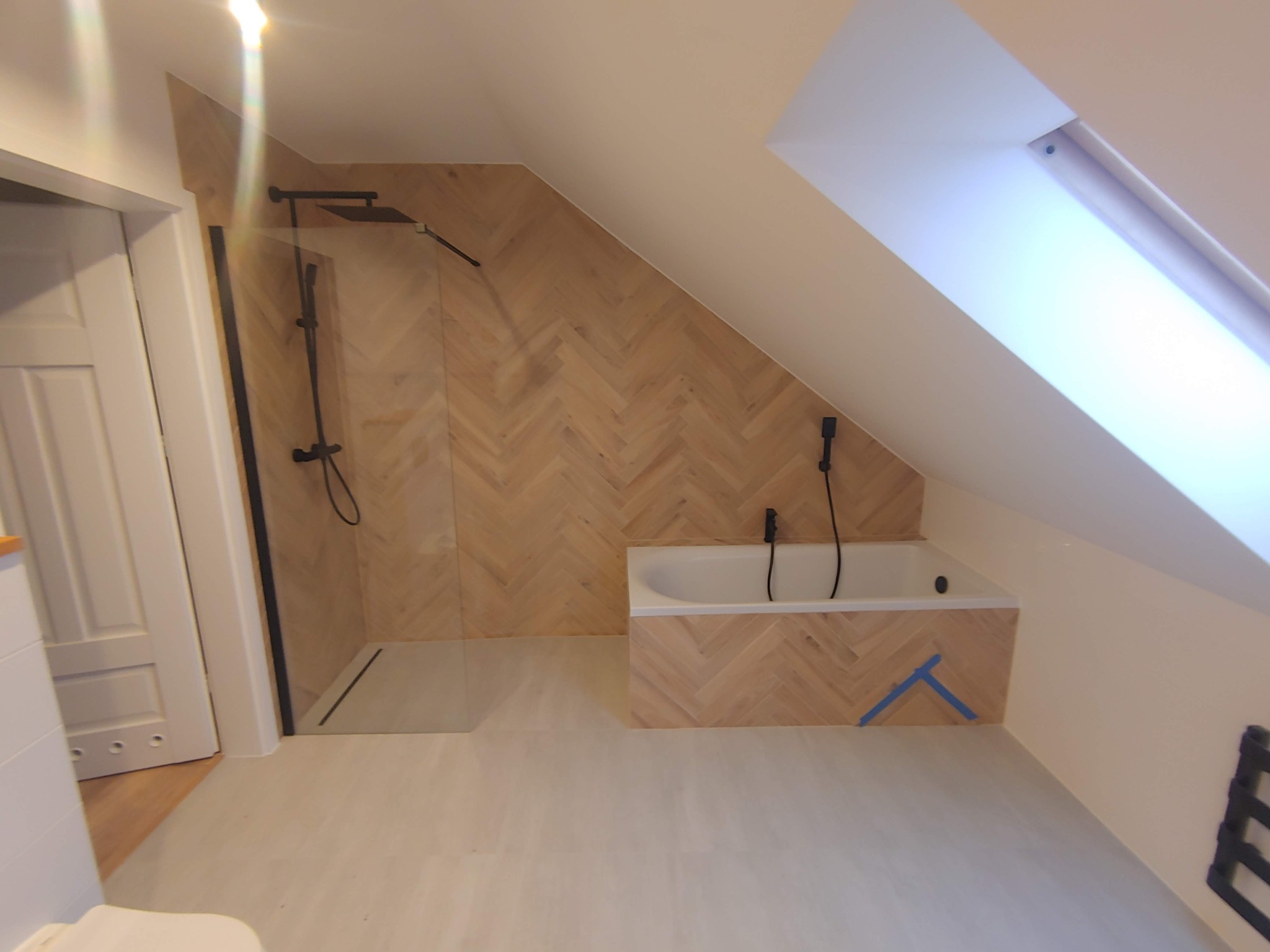 Wykończona łazienka na poddaszu ze skośnym oknem, kabiną prysznicową typu walk-in z czarną armaturą i wanną obudowaną płytkami ułożonymi w jodełkę, zabezpieczona niebieską taśmą malarską.