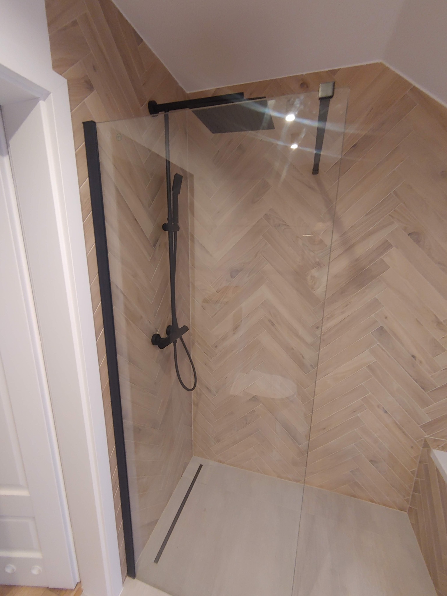 Kabina prysznicowa z czarną armaturą i szklanymi drzwiami na tle ściany wyłożonej płytkami w jodełkę, widok z boku.