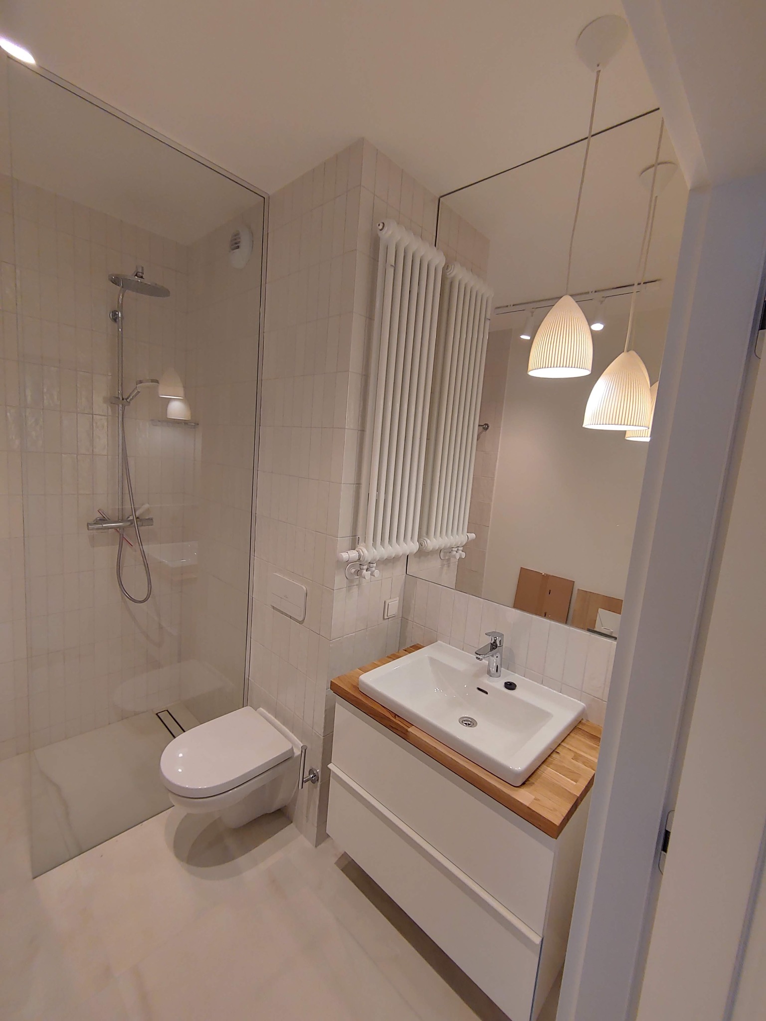 Nowoczesna łazienka z kabiną prysznicową typu walk-in, podwieszaną toaletą, umywalką nablatową na drewnianym blacie i białym grzejnikiem drabinkowym, oświetlona trzema wiszącymi lampami...