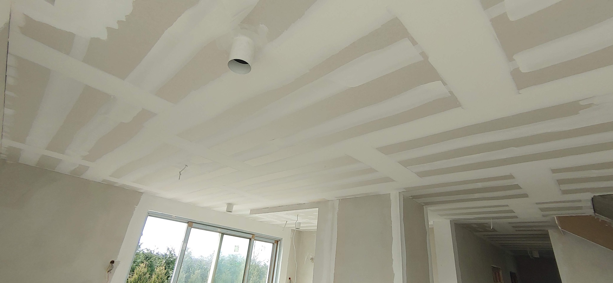 Sufit w trakcie szpachlowania z widocznymi łączeniami płyt gipsowo-kartonowych pokrytymi warstwą szpachli, z zamontowanym punktem oświetleniowym.