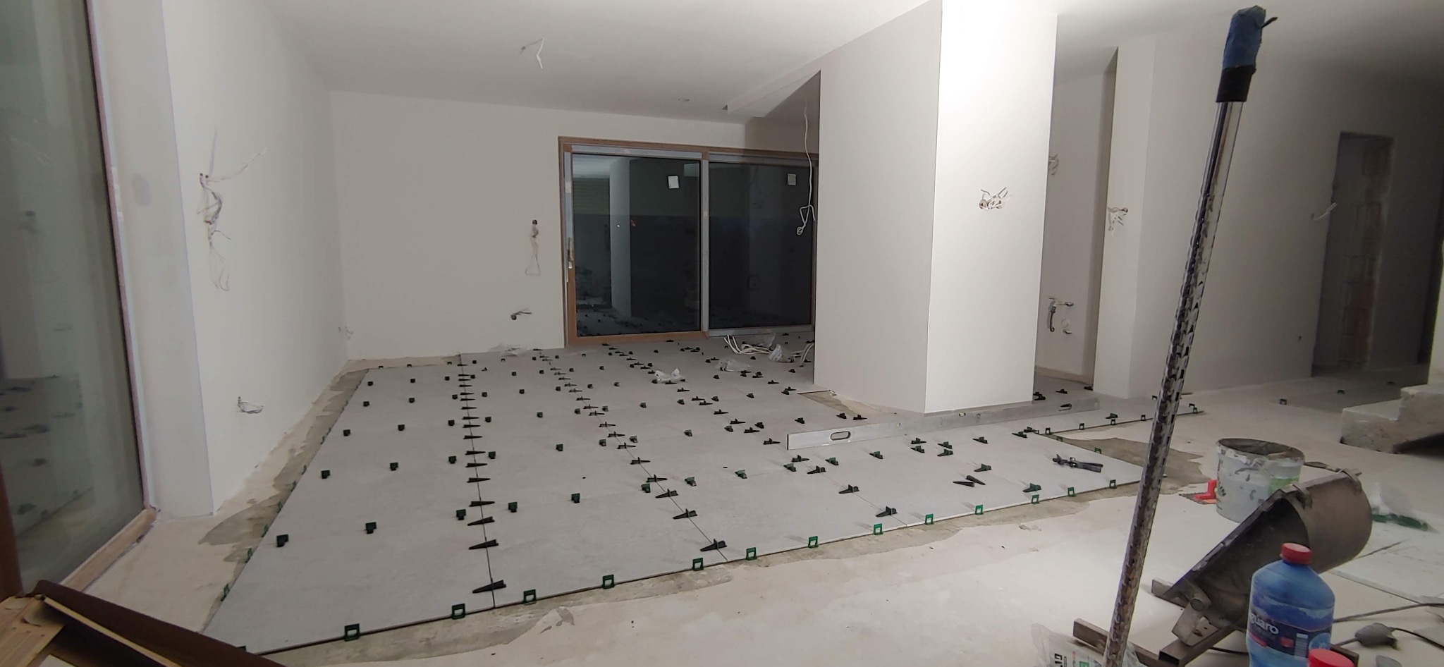 Montaż płytek podłogowych w jasnym pomieszczeniu, widoczne elementy poziomujące, narzędzia i niedokończone instalacje elektryczne.