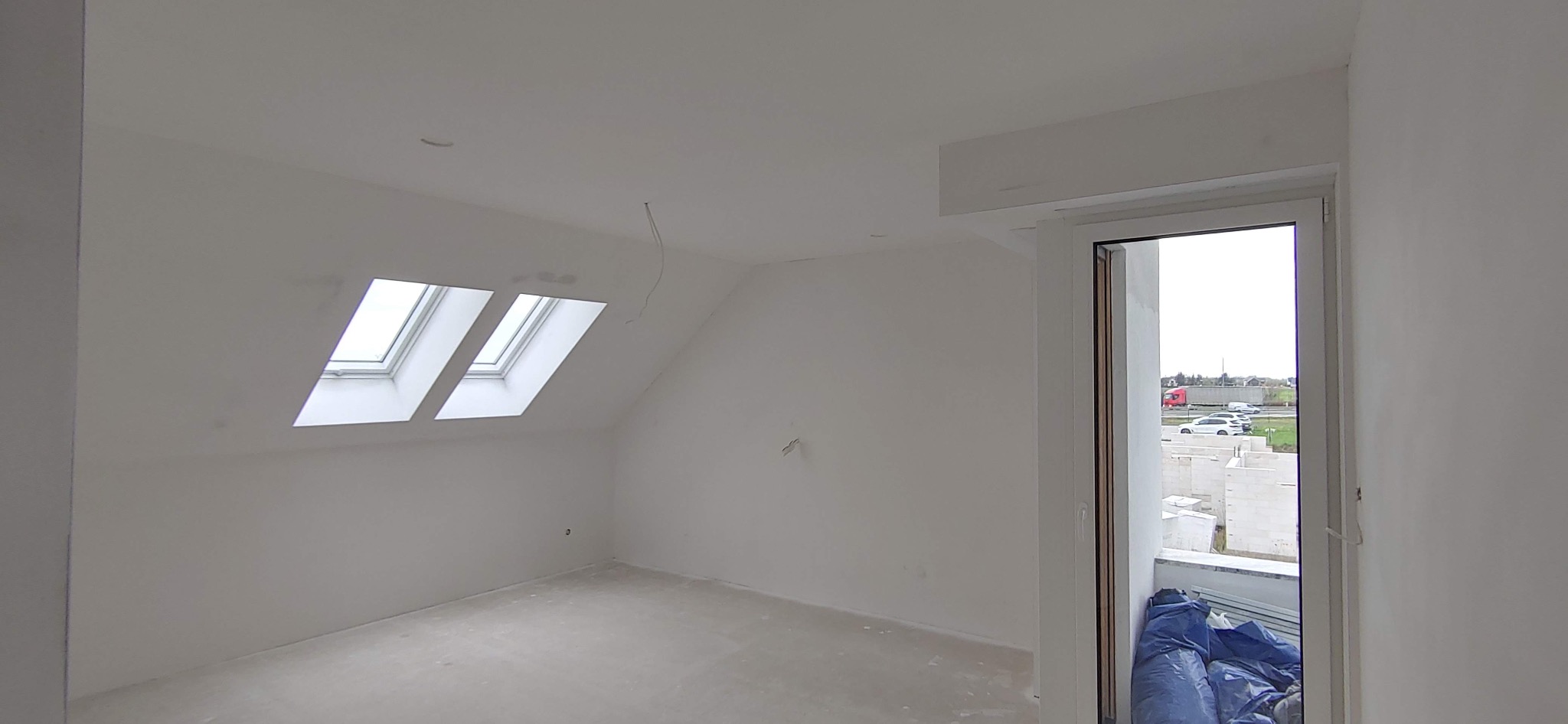 Poddasze po szpachlowaniu i malowaniu na biało, z trzema oknami dachowymi i widocznym przewodem elektrycznym zwisającym z sufitu, przez otwarte drzwi balkonowe widać plac budowy.