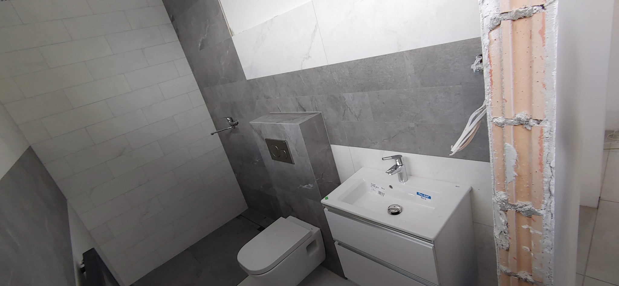 Nowoczesna łazienka w trakcie remontu, widoczna umywalka z szafką, wisząca toaleta, szare i białe płytki na ścianach oraz fragment ściany z odsłoniętą cegłą i przewodami.