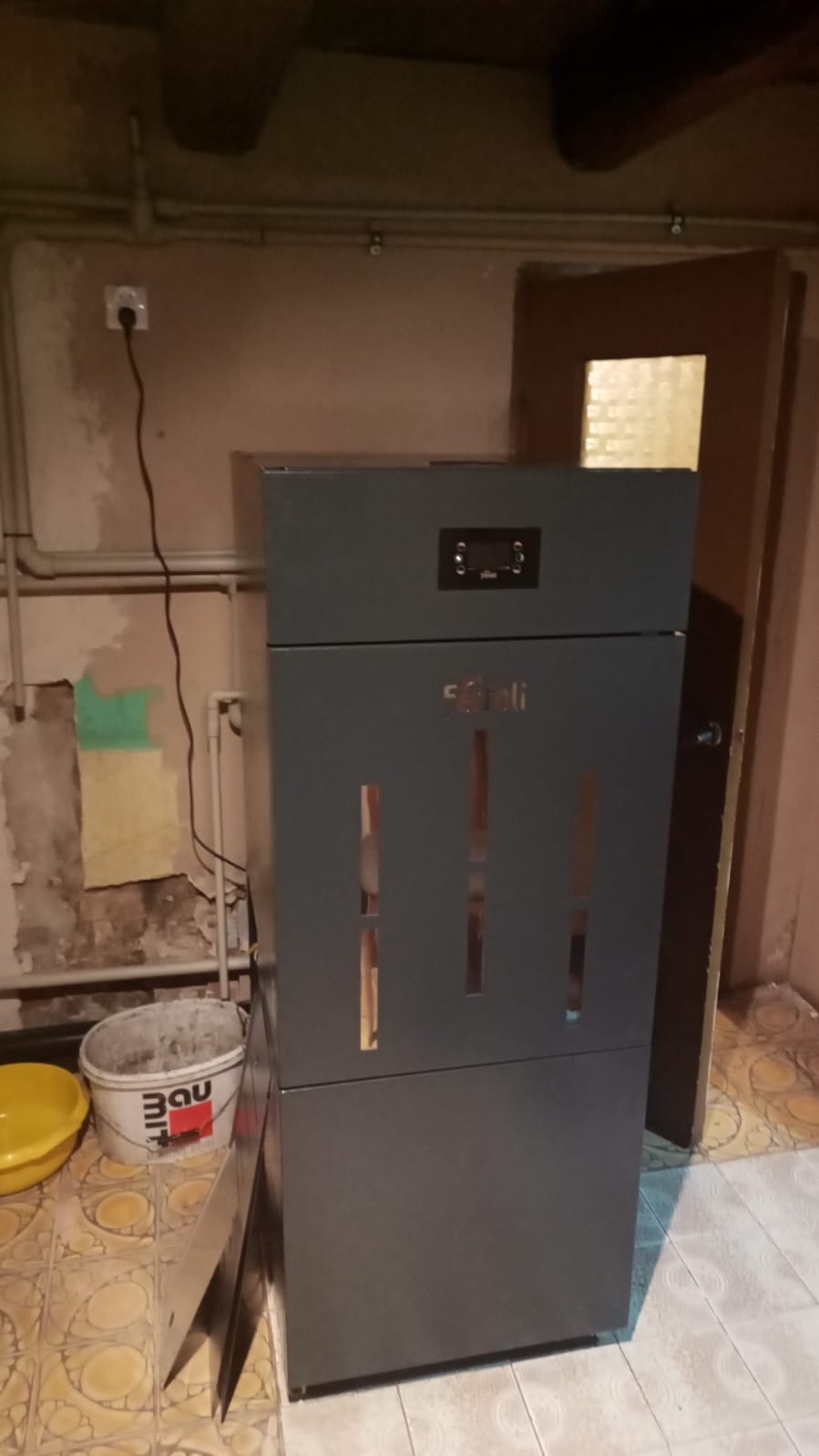 Szary piec na paliwo stałe marki Stelii z panelem sterowania, ustawiony w pomieszczeniu z niedokończonymi ścianami i ceramiczną podłogą. Obok widoczne wiadro z farbą budowlaną.