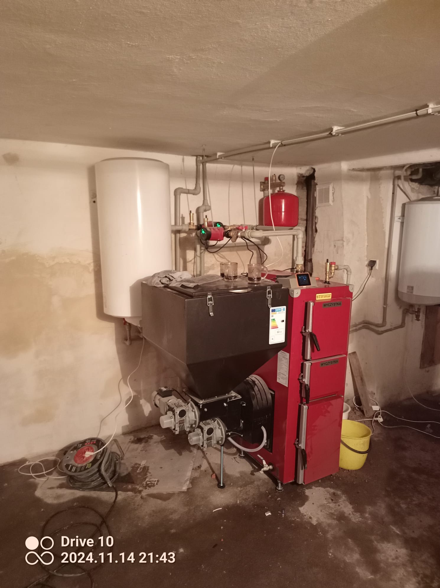 Czerwony piec na paliwo stałe z podajnikiem, widoczne elementy instalacji CO, zbiornik wyrównawczy, bojler, instalacja w piwnicy, na podłodze zwinięty przedłużacz.