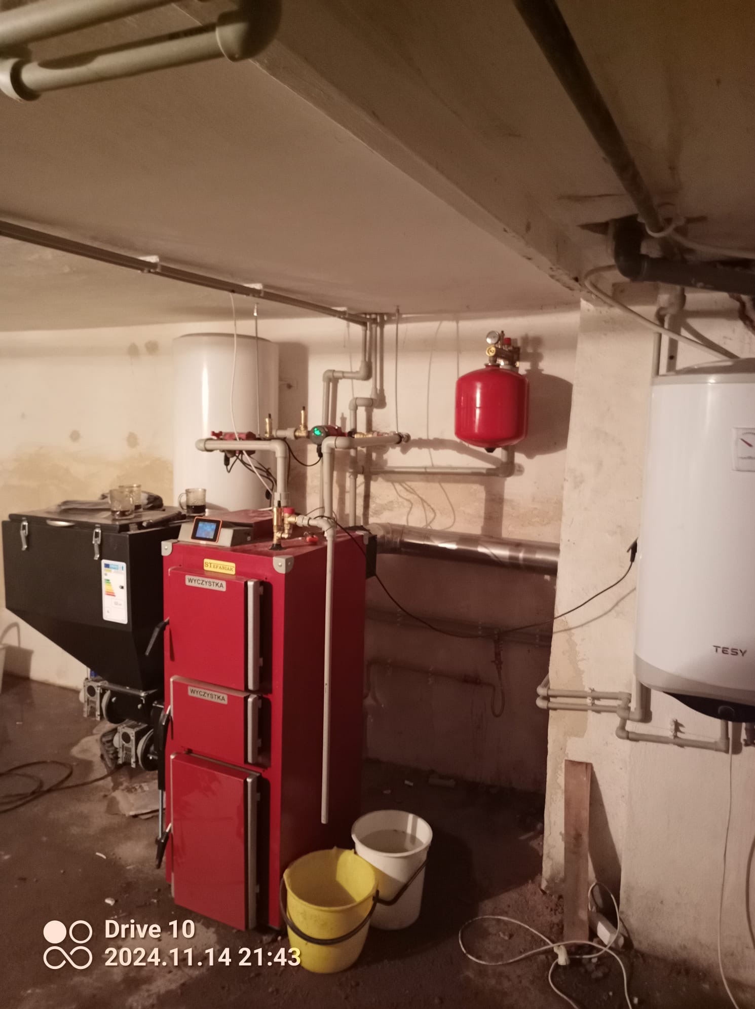 Czerwony piec na paliwo stałe z podajnikiem, widoczne rury instalacji CO, zbiornik wyrównawczy, zasobnik na paliwo, dwa wiadra obok pieca, piwniczne otoczenie.