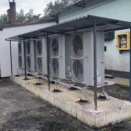 BPOWER SP.Z O.O - Rząd zewnętrznych jednostek klimatyzacyjnych pod metalowym zadaszeniem, zamontowanych na betonowym fundamencie z regulowanymi wspornikami, obok budynku z żółtą skrzynką elektryczną na ścianie.