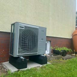 BPOWER SP.Z O.O - Szara jednostka zewnętrzna klimatyzatora zamontowana na betonowej podstawie i czarnych podkładkach, ustawiona przy ceglanej ścianie budynku z jasną elewacją i dużą, glinianą amforą w tle.