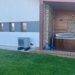 BPOWER SP.Z O.O - Szara pompa ciepła powietrze-woda zamontowana na czarnych podstawach przed biało-szarą ścianą domu z brązowymi oknami, obok drewniany taras z jacuzzi i mopem.