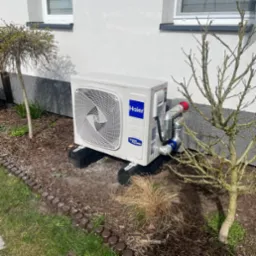 Biała jednostka zewnętrzna klimatyzacji Haier zamontowana na czarnych podstawach, z widocznymi rurami w srebrnej izolacji i czerwonymi zaślepkami, ustawiona na ziemi obok ściany budynku i małego...