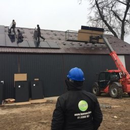 BPOWER SP.Z O.O - Montaż paneli słonecznych na dachu budynku gospodarczego, widoczny podnośnik teleskopowy i pracownicy w trakcie instalacji, w tle pracownik w niebieskim kasku z logo firmy na kurtce.