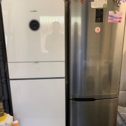 BPOWER SP.Z O.O - Biała pompa ciepła marki Bosch zamontowana obok srebrnej lodówki z magnesami i kartkami, widoczne rury instalacyjne.