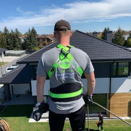 BPOWER SP.Z O.O - Instalator z tyłu, w uprzęży bezpieczeństwa z logiem firmy, trzymający wiertarkę akumulatorową na dachu domu z panelami fotowoltaicznymi w tle.