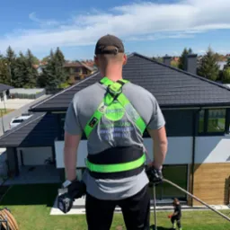 Instalator z tyłu, w uprzęży bezpieczeństwa z logiem firmy, trzymający wiertarkę akumulatorową na dachu domu z panelami fotowoltaicznymi w tle.