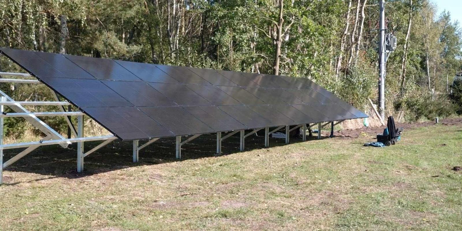 Czarny panel słoneczny na metalowym stelażu w trawiastym terenie, z lasem w tle i skrzynką narzędziową obok.