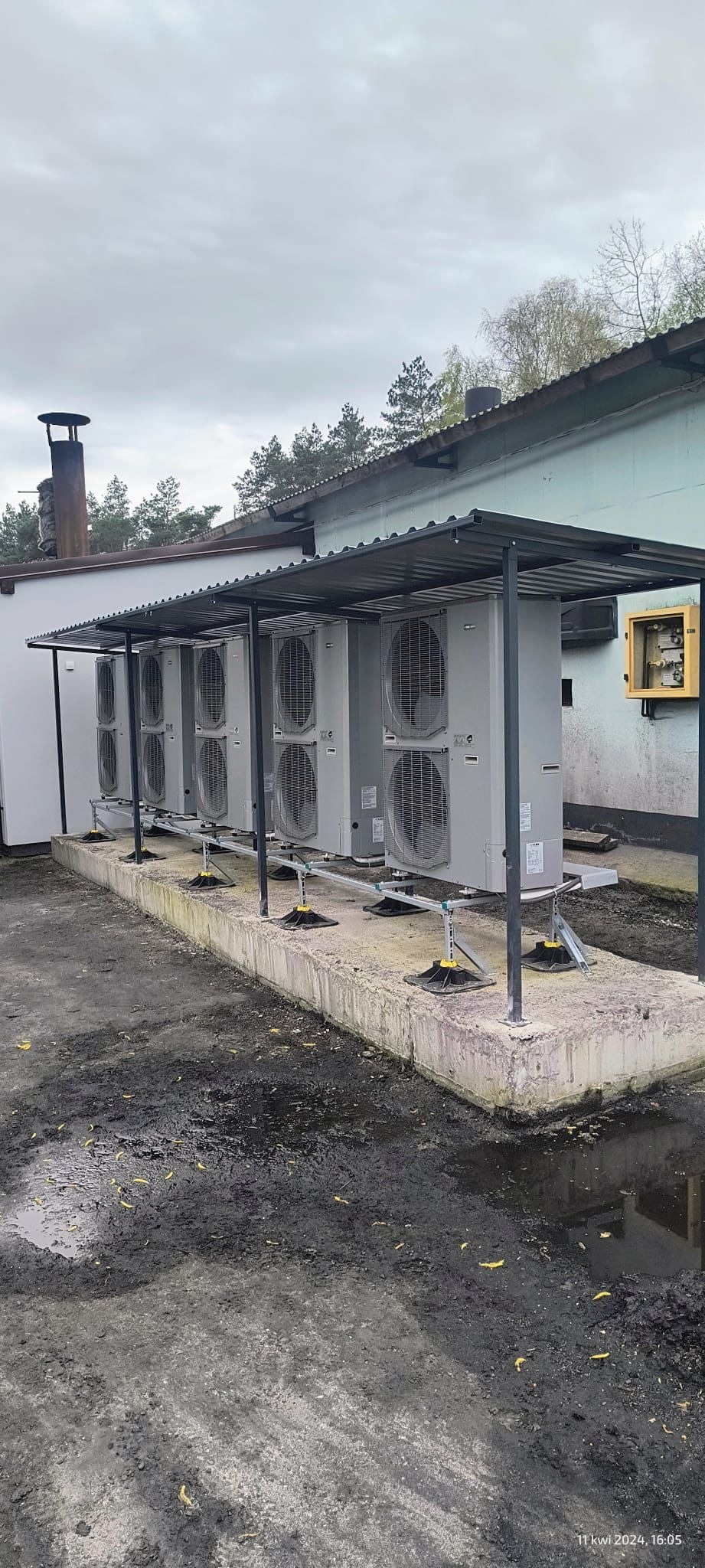 Rząd zewnętrznych jednostek klimatyzacyjnych pod metalowym zadaszeniem, zamontowanych na betonowym fundamencie z regulowanymi wspornikami, obok budynku z żółtą skrzynką elektryczną na ścianie.