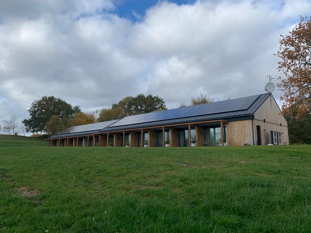 Długi, parterowy budynek z drewnianą elewacją i panelami słonecznymi na dachu, otoczony zielonym trawnikiem i drzewami pod pochmurnym niebem.