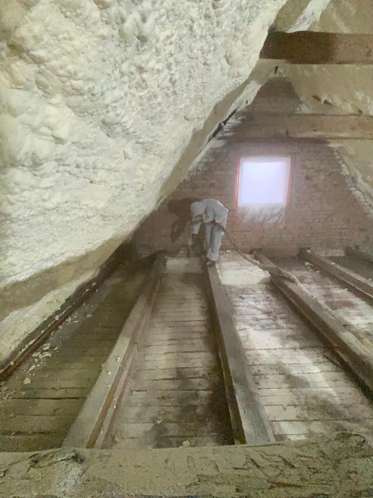 Pracownik w kombinezonie ochronnym natryskuje pianę izolacyjną na poddaszu z drewnianą konstrukcją i ceglaną ścianą z oknem.