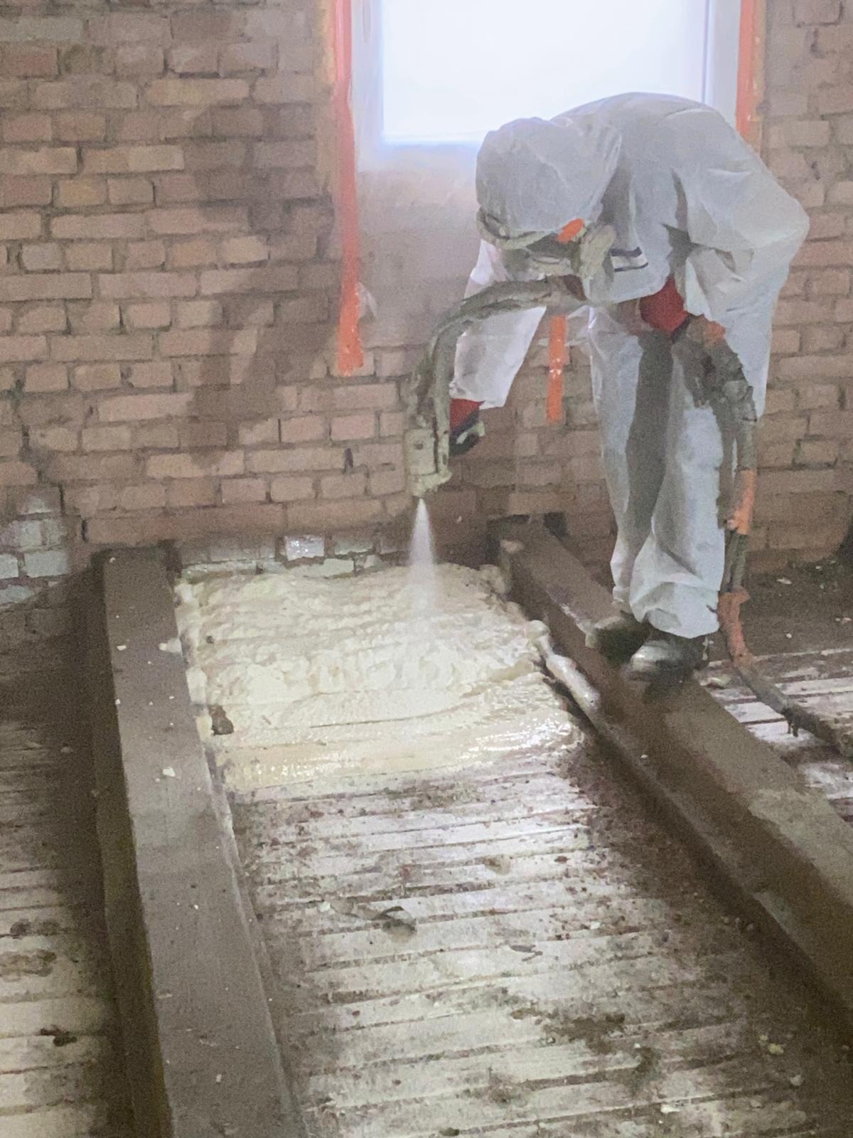 Pracownik w kombinezonie ochronnym aplikuje pianę izolacyjną na drewnianą podłogę poddasza w budynku z ceglanymi ścianami.