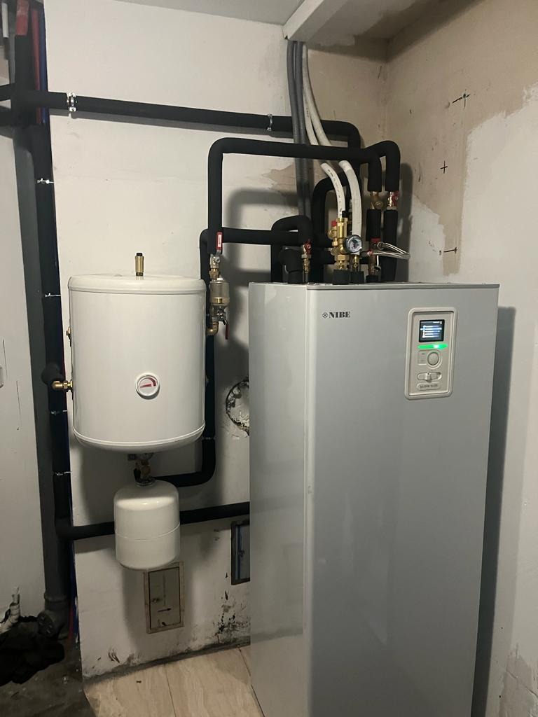 Instalacja pompy ciepła NIBE z zasobnikiem ciepłej wody i rurami w piwnicy z surowymi ścianami.