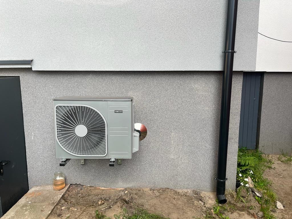 Szara zewnętrzna jednostka klimatyzacji zamontowana na elewacji budynku z szarą strukturą, obok pionowa rura spustowa, w tle widoczny fragment drzwi i roślinność.