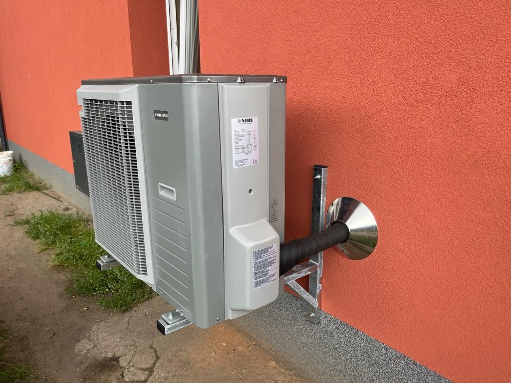 Zewnętrzna jednostka klimatyzacji NIBE zamontowana na ścianie budynku o pomarańczowej elewacji z widocznym elementem odprowadzającym skropliny i częścią instalacji.