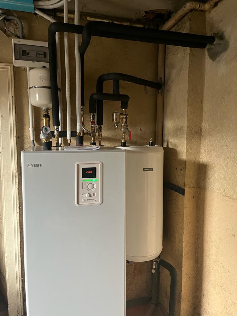 Biała pompa ciepła Nibe z podłączonym zasobnikiem ciepłej wody i czarnymi rurami izolowanymi, zamontowana przy ścianie z widoczną instalacją elektryczną.
